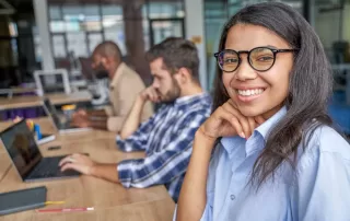 Women with glasses smiling at her desk with colleagues working in the background