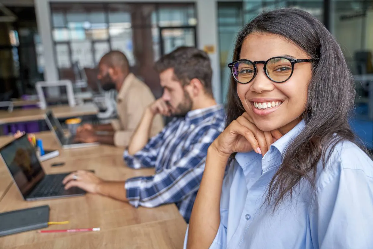 Women with glasses smiling at her desk with colleagues working in the background
